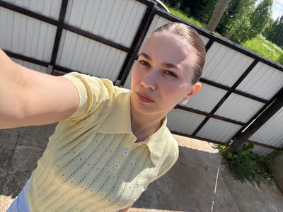 A young woman is depicted in a close-up, high-angle selfie taken outdoors in Nizhnyaya Gar', Russia. She has light skin, dark eyebrows, and her dark hair is slicked back. She looks directly at the camera with a neutral expression. She wears small, ornate earrings, possibly white pearl or gemstone with a gold-tone outline, in both earlobes. Her attire consists of a light yellow, short-sleeved collared polo shirt featuring an openwork knit pattern with vertical striped details and small eyelets. The shirt has a three-button placket with white buttons. A portion of light-colored lower garments, likely white or light blue trousers, is visible at the bottom left. The background features a white corrugated metal roof structure supported by dark brown or black beams, which appears to be a shed or an outbuilding. Behind this structure, green trees and foliage are visible under a bright blue sky with sparse clouds, indicative of a sunny day. The ground in the foreground and beneath the structure is concrete with visible cracks and patches of weeds, including dandelions, growing at the base of the structure. Direct sunlight casts shadows across the scene.