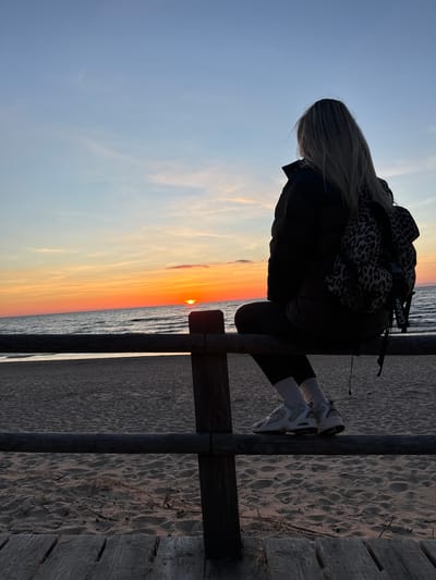 A woman with long blond hair sits on a wooden fence at the beach in Kalngale, Latvia, facing the Baltic Sea and watching the sunset. She wears a black jacket and white sneakers, and a leopard-print scarf or shawl is draped over her shoulders. The sunset creates an orange and yellow gradient in the sky and reflects on the water. Sand covers the beach area near the fence.