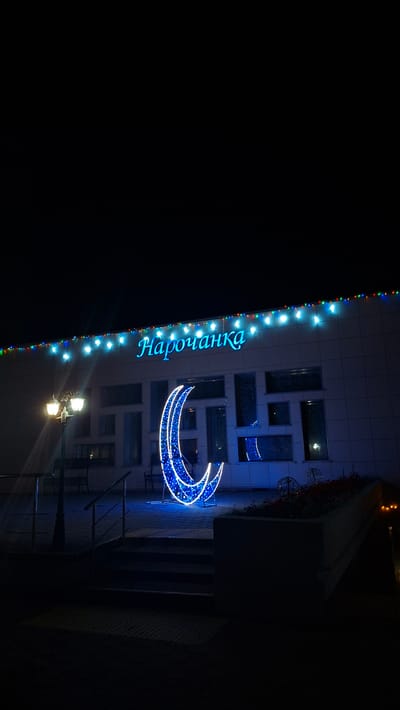 At night in Нарацкі сельскі Савет, Belarus, a building is decorated with blue lights outlining the word "Нарочанка" and a crescent moon. The building has a modern facade with rectangular windows. String lights run along the building's edge. A streetlight illuminates the area. Stairs are visible to the right of the illuminated crescent. The background is dark.