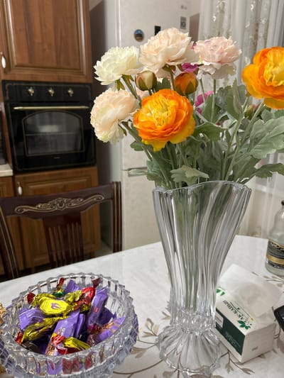 This is a forensic, matter-of-fact description of an indoor scene, likely a kitchen or dining area, located in Нижнее Казанище, Russia. The image shows a table covered with a tablecloth, on which are placed a crystal bowl filled with individually wrapped chocolates, a clear glass vase containing a bouquet of artificial flowers, and a box of tissues.
The chocolates are a mix of purple and yellow wrappers. The bouquet consists of flowers in shades of orange, yellow, and pale pink, with green artificial foliage. The vase is tall and clear, with a ribbed, fluted design. The box of tissues has green text and imagery.
In the background, a wooden kitchen cabinet with a built-in oven is visible. The oven has a dark glass door and several control knobs. To the right of the vase, a portion of a refrigerator is visible, along with a white curtain. The overall environment appears to be a domestic setting. No people are present in the image.
