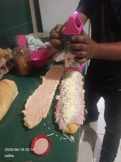 A person, whose hands are visible, is grating cheese onto a sandwich. The sandwich is on a green surface, which appears to be a table or countertop. Part of a loaf of bread, spread with a pink substance, is to the left of the sandwich. To the right of the sandwich, a person's legs, dressed in black pants, are visible. In the background, there is a pink bowl, a bottle with contents, and what appears to be a white bag with black markings. The scene is dimly lit. The image includes a timestamp and a name overlay: "2026/06/18 20:14 natha". The location is stated to be El Chaparral, Venezuela.