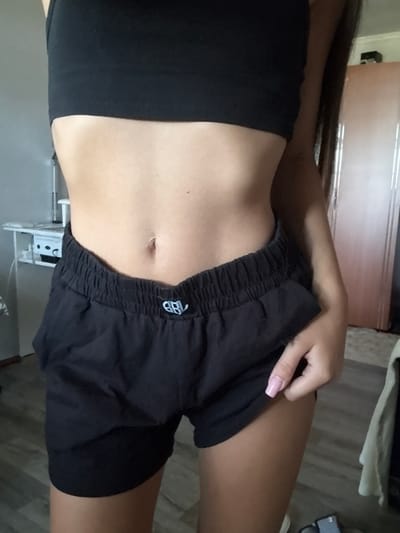 The image shows a close-up of a person's midsection, from the navel down to the upper thighs. They are wearing a black sports bra and black shorts with a cinched waist. The background includes a dresser or wardrobe, a portion of a wall, and a sliver of a floor. The scene appears to be indoors, likely in a residential setting. There are no recognizable landmarks or elements that would place this image specifically in Basarabeasca, Moldova.