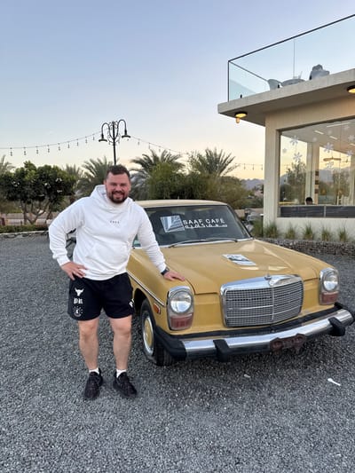 An adult male with a light complexion, dark hair, and a beard stands smiling in an outdoor setting at SAAF CAFE in Hatta, United Arab Emirates. He is positioned to the left of a vintage yellow Mercedes-Benz sedan, with his left hand on his hip and his right hand resting on the car's front fender. He wears a white hooded sweatshirt, black athletic shorts with white "PR" and bull head logos, and black athletic shoes. The car's windshield features a 'SAAF CAFE' sticker in English and Arabic. The scene is set on a grey gravel surface with a modern two-story building, identified as SAAF CAFE, on the right, featuring large glass windows with snowflake decals and an upper-level outdoor seating area with a glass railing. Distant palm trees, vegetation, a decorative street lamp with string lights, and mountains under a twilight sky complete the background.