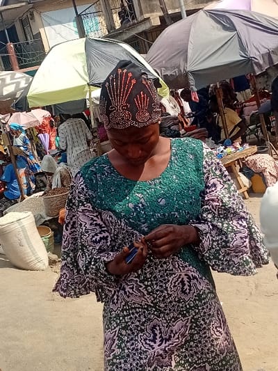 The image depicts an outdoor market scene in Ogaminan, Nigeria. In the foreground, an adult female with dark skin is central, looking downwards. She wears a long-sleeved dress featuring a green, purple, and white patterned fabric, and a black headwrap adorned with symmetrical golden-orange beadwork in a fan-like design. Her hands are positioned near her chest; she holds a small blue object in her left hand and is actively manipulating a small, thin object with her right hand.
The background reveals a bustling market environment with numerous individuals, most partially obscured. Multiple large umbrellas provide shade, including a prominent black and light green umbrella directly behind the primary subject. Market stalls with various goods are visible, and a large white sack, likely containing bulk items, rests on the ground to the left. Woven baskets are discernible among the merchandise. Buildings are visible in the distant background, and the ground appears to be a paved surface.