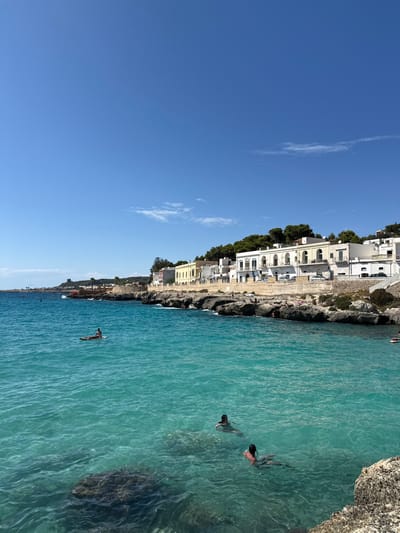 The image displays a coastal scene in Santa Maria al Bagno, a hamlet within the city of Nardò, Italy. The sky is clear and bright blue, with minimal, wispy white clouds present in the upper right. The foreground and mid-ground are dominated by clear, turquoise-to-blue seawater, through which dark, irregular rock formations are visible on the seabed.

Three individuals are in the water. One person is positioned prone on a light-colored paddleboard in the mid-left portion of the water. Two other individuals are partially submerged, swimming or wading in the immediate foreground, with their backs generally facing the viewer. On the distant rocky shoreline to the left, several small, indistinct figures are visible, indicating the presence of other people.

The coastline, extending from the mid-ground to the background, consists of dark, rugged rock formations that meet the water. Above this shoreline, a series of multi-story buildings, predominantly white and light-yellow, are constructed along an elevated slope. These structures feature arched windows, balconies, and some have red-tiled roofs. Dense green vegetation, primarily trees resembling pines, covers the higher ground behind the buildings.

In the far distance on the left coastline, a dark, stone watchtower-like structure is visible. A small red buoy or marker floats in the water near this distant tower. Several parked vehicles are discernible on a paved area or road situated behind the buildings in the mid-ground. The scene is illuminated by strong daylight.