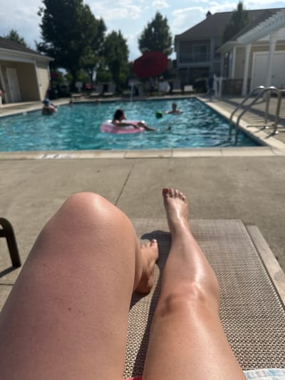 A first-person perspective shot shows a person's legs and feet resting on a lounge chair by a swimming pool. In the background, several people are swimming and playing in the pool. A pink float is visible in the water. Buildings and trees are in the far background. The location is Allen Township, United States of America.
