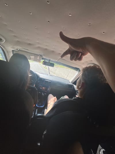 The image shows the interior of a car, likely on a road trip. The perspective is from the back seat looking forward. Two people are visible, one driving and one in the passenger seat. The driver has long, dark hair and is wearing a black shirt. The passenger has curly brown hair and is also wearing a black shirt. A hand is visible in the foreground, pointing upwards towards the car's ceiling, which is covered in numerous small circular indentations. The dashboard and a rearview mirror are visible. The road outside the car is visible through the windshield, appearing to be a paved road with concrete barriers on the sides, suggesting a mountainous or elevated terrain. The lighting indicates it is daytime, with sunlight entering the car. The scene does not contain any specific elements that would identify it as being in Leshtak, Bulgaria.