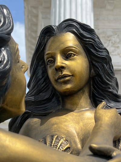 The image is a close-up, waist-up shot of a golden statue of two figures, likely mermaids, interacting. The figure on the right, facing forward, is a woman with long, black, wavy hair. Her face is rendered in a detailed, lifelike manner with distinct eyes, nose, and lips. She has a shell detail on her chest. Her left arm is raised, with her hand resting near her face. The figure on the left is partially visible, with its head turned towards the central figure, appearing to be in conversation or close proximity. This figure is also gold and has dark hair. The background consists of a bright blue sky with some white clouds and large white columns, suggesting an outdoor setting possibly near a building with classical architecture. The overall impression is one of intimacy and beauty. This statue is located at the Mermaid Statue, Thepkrasattri Road, Pa Khlok, Thalang District, Phuket, Thailand.