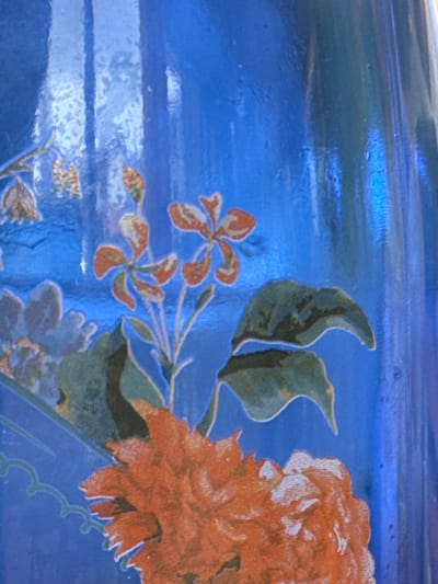 This image depicts a close-up, static view of a decorative surface, likely ceramic, glass, or a similar glazed material, located in Anzola dell'Emilia, Italy. The dominant background color is a vibrant, reflective blue, exhibiting vertical striations and minor textural irregularities. A multi-component floral motif is rendered across the surface. In the lower-center, a large, multi-petaled flower, possibly a peony or rose, is depicted in shades of red and orange with discernible textural details. Above this, slightly to the right, a stem with several four-petaled flowers, also in orange-red, is present, outlined in a lighter blue and accompanied by dark green leaves. Additional muted floral elements in bluish-grey and brown-yellow tones are visible in the upper-left and middle-left areas. No people, specific actions, or further environmental details are present.