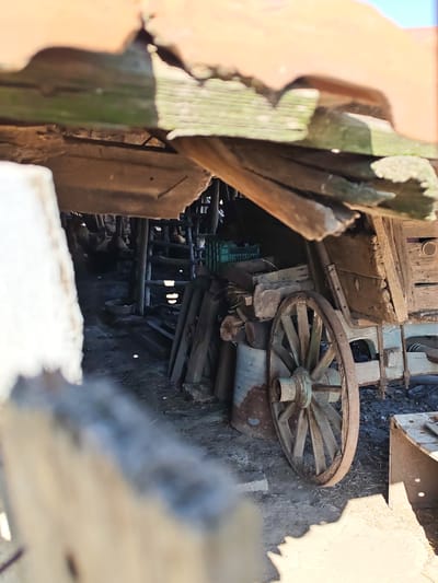 The image shows a dimly lit, cluttered interior of what appears to be a dilapidated shed or barn. The foreground is dominated by a weathered wooden wagon wheel with spokes. Behind the wheel, various wooden planks, crates, and what seems to be part of a wooden structure are visible. The background reveals a darker area with more wooden supports and possibly some chickens, evidenced by the shapes and what could be a ladder-like structure. The roof above is partially visible, made of corrugated material and decaying wood. The overall impression is one of neglect and disuse. The location is identified as Byal Izvor, Bulgaria.