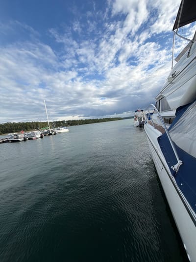 The image depicts a cloudy sky over a body of water, likely a bay or lake, with several boats moored. In the foreground, the starboard side of a white motor yacht is visible, partially covered by a blue tarp. The yacht has a chrome railing and a teak deck. In the midground, a fleet of smaller boats, including a sailboat, are docked at a pier on the left. The distant shoreline is lined with dense evergreen trees. The water is dark and exhibits small ripples, suggesting a gentle breeze. The location is identified as Molodezhnyi, Russia. There are no people visible in the image.
