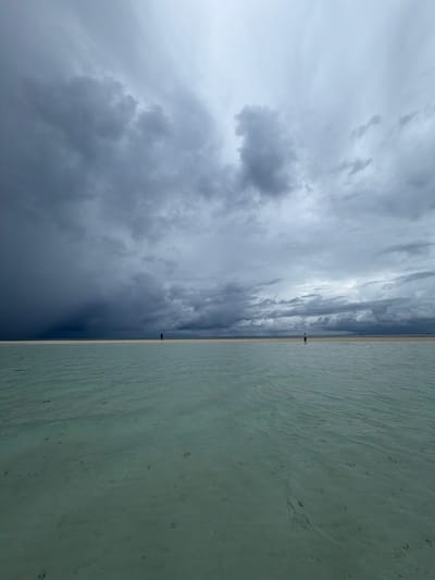The image shows a beach scene likely in Maafushi, Maldives, characterized by a distinct division between a turbulent, cloudy sky on the left and calm, shallow water on the right. A thin strip of sand separates the sky and the water. Two small figures are visible along this strip of sand. The sky is dominated by dark, heavy clouds, suggesting an impending storm. The water appears shallow with a clear bottom.