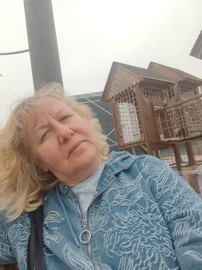 A woman with blonde, wavy hair is the subject of this medium-shot selfie, set against the backdrop of a children's playground. She is wearing a light blue zip-up jacket with a white patterned design.
Behind her, a complex wooden structure designed for children to climb and play on is visible. The structure is multi-tiered and features netted areas and platforms.
In the background, there is a modern building with glass panels, indicating the location is urban. The sky is overcast, lending a flat, muted light to the scene.
Based on the location, this photograph was taken at a playground in the city of Tver, Russia.