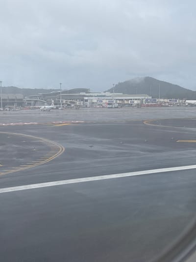 The image depicts an airport tarmac on a cloudy, overcast day, likely at Phuket International Airport (HKT) in Sakhu, Thailand. The tarmac is wet, indicating recent rain, with visible runway markings and yellow taxiway lines. Several aircraft are parked at the terminal gates, connected by jet bridges. In the background, the airport terminal building is visible with its modern architectural design. Behind the terminal, a lush green mountain range with clouds hovering around its peak is prominent, characteristic of Phuket's landscape. The overall atmosphere is subdued due to the weather conditions.