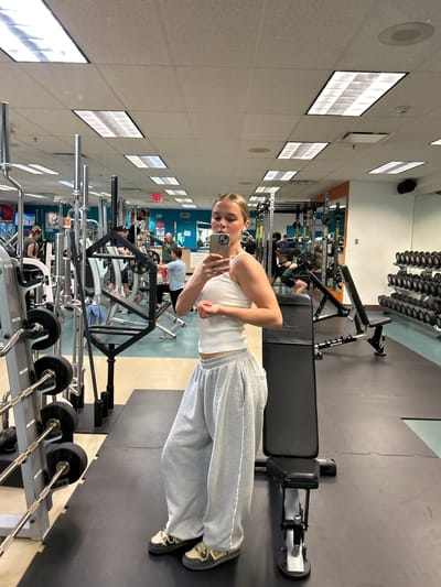 A young woman is taking a selfie in a gym. She is wearing a white tank top and grey sweatpants. She is standing in front of a weight bench. In the background, there are other people working out and various exercise machines and weights. The gym has a modern, clean design with fluorescent lighting and mirrors. The location is likely a fitness center in Whistler, Canada, given the presence of fitness equipment and people engaged in exercise.