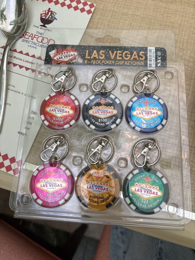 The image shows a Las Vegas 6-pack poker chip keychain set still in its clear plastic packaging. The keychains are in six different colors: blue, green, black, gold, red, and pink. Each chip has the phrase "Welcome to Fabulous Las Vegas, Nevada" on it, along with a different dollar value ($25, $100, $500, $1000, $5000, $1,000,000). The packaging has the words "LAS VEGAS 6-PACK POKER CHIP KEYCHAIN" in bold text. A spoon and a menu for "THE SEAFOOD SPECTACULAR" buffet can be seen partially in the bottom left of the frame. The location is indicated by the Las Vegas memorabilia, but not specifically the city of Winchester.