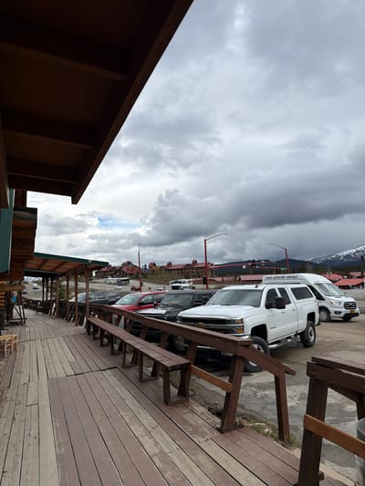 The image depicts a view from a raised wooden deck in Denali Park, United States, showing parked vehicles and buildings under a cloudy sky.

The foreground features a section of a brown wooden deck with visible planks and supporting beams. In the midground, a row of parked vehicles, including white vans and a red car, lines a street. Buildings with red roofs are visible behind the vehicles, and traffic lights are present at an intersection. In the background, snow-capped mountains are visible. The sky is overcast with dark, heavy clouds dominating the upper portion of the image.