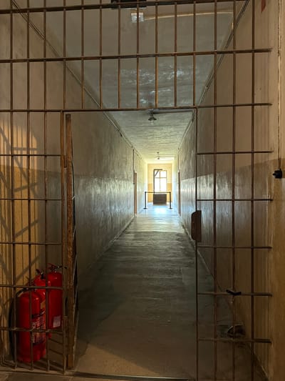 This image shows an upward view of a narrow, tall interior space, possibly a ventilation shaft or a confined stairwell, with metal grating in the foreground. The walls are rough and textured, illuminated by light filtering from a window at the top. The scene appears to be indoors, with a stark, utilitarian aesthetic.
Prominently featured in the lower right of the frame, behind the grating, are two red fire extinguishers. They are cylindrical and appear to be standard fire safety equipment. Above the fire extinguishers, there's a wooden plank or beam, also behind the grating, and further up, a dark object is affixed to the wall.
The metal grating is a recurring element, creating a layered effect and suggesting a secure or enclosed environment. Sunlight streams in from the square window at the very top, creating patterns of light and shadow on the walls. The overall impression is one of confinement and a focus on infrastructure rather than human presence. The image lacks any indicators of time of day or specific weather conditions, but the enclosed nature of the space suggests it is within a building. The location context of Oświęcim, Poland, and the stark, potentially historical architecture hinted at by the raw materials and functional design, might evoke associations with such sites. No discernible text is visible on the walls or objects, except for labels on the fire extinguishers which are too blurry to read.