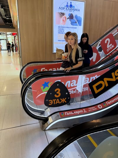 The image shows an escalator in what appears to be a shopping mall. Several individuals are visible on the escalator, some of whom are looking at their phones. A young woman in the foreground is holding cups and looking towards the camera. Red signage with Russian text is present on the side of the escalator, indicating floor numbers and store names, including "2 этаж" (2nd floor) and "Галамарт" (Galamart), and "DNS". In the background, a sign for a medical clinic ("ЛОР КЛИНИКА" - ENT CLINIC) is visible. The overall environment suggests an indoor commercial setting in Moskovskiy, Russia.