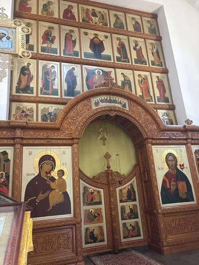 The image depicts the interior of the Church of the Iberian Mother of God in Sestroretsk, Russia. The focal point is the ornately carved wooden iconostasis, which dominates the scene. The iconostasis features multiple rows of religious icons, painted in a traditional style, depicting various saints and biblical scenes. The icons are framed by intricate wooden carvings. In the center of the iconostasis, a large arched opening reveals a gilded backdrop. Flanking this opening are two large icons; one depicts the Virgin Mary holding the Christ child, and the other shows Jesus Christ. The wooden structure extends across the frame, with various carved panels and decorative elements. The overall impression is one of a richly decorated and spiritual environment within a church.