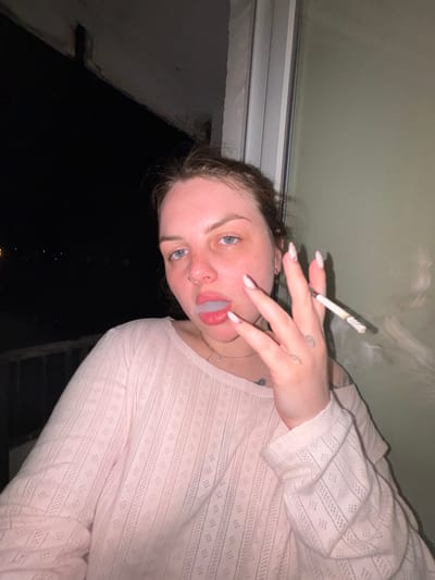 A close-up, tilted portrait features a young adult female holding a lit cigarette and exhaling a cloud of smoke from her mouth. Her face is directed towards the camera, revealing light blue eyes, a nose ring, and subtle freckles. Her lips are slightly parted, forming a shape as the smoke exits. Her hair, partially visible, appears dark brown.

She is wearing a light pink, long-sleeved top with a distinctive patterned or open-knit texture. Her nails are manicured with light polish and delicate designs. Small tattoos are visible on her fingers and the back of her hand, and a thin necklace adorns her neck.

The background above her consists of a plain, light-colored wall or ceiling, while the area behind and to her left, seen through what appears to be a window or from a balcony, is dark, indicating nighttime. Faint distant lights are visible in the dark background. A window frame or structural element is present on the left side of the image.

The activity depicted is smoking. The overall impression is a casual, nighttime moment. There is no visible text in the image. The provided location context for this media file is Chornomorskoe, Ukraine.