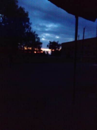 The image depicts a dark, blurry outdoor scene, likely taken at dusk or twilight. A dark foreground occupies the lower half of the frame, suggesting a ground surface such as pavement or dirt. In the midground, silhouetted trees with dense foliage are visible against a twilight sky. The sky is a deep blue with hints of lighter hues near the horizon, where a faint glow suggests the setting sun or distant lights. In the background, indistinct shapes of buildings or structures are present, one appearing to be a long, low building and another a taller, darker structure. A vertical pole or support is also visible against the sky. The overall impression is one of a dimly lit, possibly rural or suburban, environment at the end of the day. Without further information or sharper detail, pinpointing the exact location within Rímac, Peru, is not possible based solely on this image.