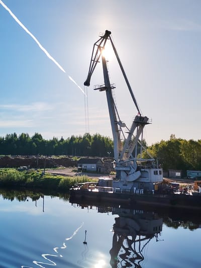 This vertical photograph captures an industrial scene along a calm waterway at Annensky Most, Russia, under a clear, bright sky. The dominant subject is a large, grey gantry crane, likely a portal or level luffing type, mounted on a barge or pontoon. The crane is heavily backlit by a low-angle sun, which creates a prominent lens flare behind its complex lattice structure, partially silhouetting it.
The sky is a vibrant blue, featuring a long, wavy white contrail from an airplane stretching diagonally from the top-left to the bottom-right. The tranquil water of the river or canal perfectly mirrors the blue sky, the brilliant sun, the crane, and the contrail, with subtle ripples adding texture to the reflections.
The far bank of the waterway is lined with lush green vegetation, which gives way to a dense forest of trees in the background. On the land directly behind the crane, there are extensive piles of logs or timber, suggesting an active timber yard or logging facility. Several small, utilitarian buildings, possibly shacks or offices, are visible among the timber, along with a utility pole and an old, dark vehicle.
The time of day appears to be either early morning or late afternoon, given the low position of the sun and the resulting backlighting. The scene conveys a sense of stillness and quiet, highlighting the juxtaposition of industrial infrastructure with the natural environment. There is no visible human activity, and no legible text can be discerned on any of the objects.