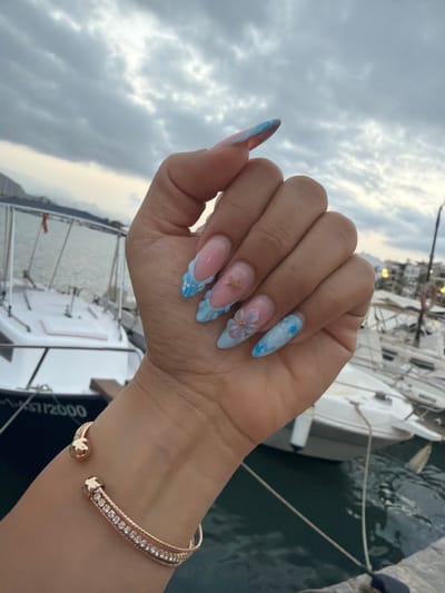 A close-up shot features a person's left hand with meticulously manicured nails against a backdrop of boats docked in a marina, possibly in Port D'alcúdia, Spain. The nails are long and almond-shaped, decorated with a French tip design incorporating light blue and white swirls reminiscent of clouds or waves. Some nails also feature delicate embellishments, including a small gold starfish and a translucent light blue flower. A rose gold bracelet adorned with sparkling cubic zirconia stones is worn on the wrist. The background shows several white boats with dark hulls, some with railings and windows. The water is dark and calm, and the sky above is overcast with heavy gray clouds, suggesting possibly impending weather or the late evening. The overall impression is one of leisure and sophisticated style, juxtaposed with the maritime setting.