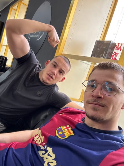 This image captures two males inside a modern barbershop in Krems an der Donau, Austria. In the foreground, on the right, a male with short light brown hair, a trimmed beard, and metal-framed glasses is positioned, holding the camera. He is wearing a blue and red FC Barcelona soccer jersey with gold lettering on the blue section. Behind him, to the left, a second male with a buzz-cut hairstyle and a mustache is seated on a dark, possibly leather, chair or couch. He is wearing a dark gray short-sleeved t-shirt and is flexing his left bicep, with his fist clenched. Both individuals are looking directly at the camera.
The background reveals typical barbershop elements: a large, dark oval sign or mirror on the left displaying "BARBER SALON" in white text alongside a barber pole graphic. To the right, a shelving unit with gold-colored vertical supports and light-colored shelves holds various retail hair products, including boxes labeled "MOWB PROFESSIONAL" and a red bag with white Japanese characters. A window, showing bright external light, is visible beyond the shelving.