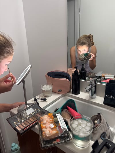 A young female is shown applying makeup in a bathroom. She is visible in the foreground, focusing on her right eye with a small brush while looking into a lighted magnifying mirror. Her reflection in the large wall mirror above the sink shows her leaning forward, performing the same action. She has light hair tied back and is wearing a gray tank top. The bathroom features white walls and a light-colored counter with a white ceramic sink.

The counter and sink area are cluttered with numerous items:
On the counter to the left, an open "NAKED" eyeshadow palette is visible, alongside Q-tips and hair ties.
Near the sink, a pink "KUSSHI" makeup bag is open. Two black pump dispensers are positioned next to a chrome single-handle faucet. A black towel embroidered with "Makeup" lies to the right of the faucet.
Within the sink basin, a teal-colored bag contains a hairbrush with pink bristles and a pink squeeze tube. Another small, grey-patterned makeup bag is also in the sink.
Two clear glasses filled with water and ice cubes are present: one on the counter to the left of the pink makeup bag, and another in the sink on the right side.
A clear plastic container of pre-packaged food, labeled "Spicy Shrimp Roll" and "SHRIMP POKE," containing cooked shrimp on rice, is placed on the counter near the sink. A black hair clip is on the far right edge of the sink.

The precise location within Manhattan Beach, United States, cannot be determined from the visual information provided.