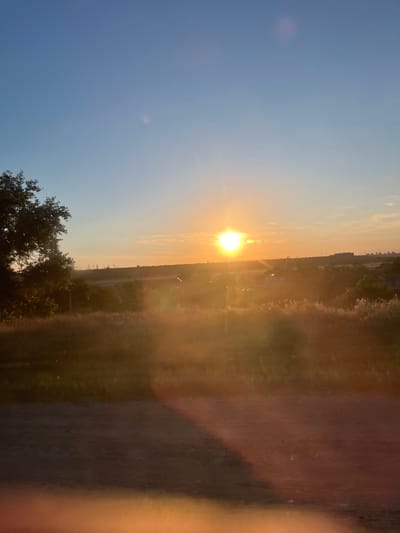 A sun setting over a rural landscape with a dirt road in the foreground. The sun is bright and low in the sky, casting a warm glow over the scene. There are trees and some buildings in the distance, as well as power lines. The sky is a clear blue with a few wispy clouds. The image was taken in Karagash, Moldova.