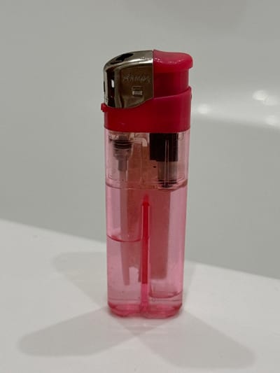 This media file features a close-up, side-view of a pink, transparent, disposable lighter. The lighter is oriented horizontally, with its metallic igniter mechanism on the left and the body extending to the right. A red plastic piece partially covers the top of the igniter. Visible within the transparent pink body are the inner workings of the lighter, including what appears to be a fuel reservoir and a metal striker. The surface beneath the lighter is plain and white, with a soft shadow cast to the right, suggesting a light source from the left. The lighting is even and diffused, indicating an indoor setting with artificial light or an overcast day. There is no discernable activity occurring, as the lighter is stationary. The image is sharp and detailed, allowing for a clear view of the lighter's construction. Faint, cursive text is visible on the metallic part of the igniter, but it is too indistinct to be read. The overall impression is a simple product shot, focusing solely on the object itself.