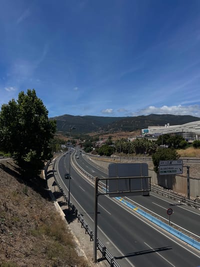 The image shows a highway curving through a landscape with mountains and some buildings. The sky is blue with some wispy clouds, suggesting it is daytime and the weather is clear. 

The highway has multiple lanes and is marked with white and yellow lines. A speed limit sign indicates 60. There are several cars on the road. On the right side of the highway, there is a blue and yellow border. Road signs are visible, one indicating "400 m" with an arrow pointing left and "ALGESIRAS costa" and "avda. Agua Marina". Another sign shows "1120" and a letter "R" in a square. 

In the background, there are green mountains with some vegetation. A large building complex with a white facade is situated at the base of the mountains, possibly a shopping center or industrial area, with a "PRIMARK" logo visible. 

In the foreground, a large, leafy green tree is prominent on the left, casting a shadow. The ground in the foreground and along the side of the highway is covered with dry vegetation and rocks. The scene appears to be of a sunny day in a coastal area of Spain, near Algeciras. No people are clearly visible.