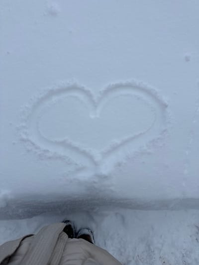 The scene depicts a top-down view of a snow-covered outdoor environment in Probuzhdenie, Russia. A distinct heart shape has been drawn into a layer of fresh white snow, appearing as a shallow indentation exposing a slightly darker layer beneath. The heart occupies the upper-mid section of the frame. In the foreground, at the bottom of the image, the feet of an individual are visible, clad in dark-colored shoes or boots, partially obscured by light-colored (beige or off-white) winter clothing, likely a coat or parka. The person stands on a lower level of snow, separated from the area containing the heart by a discernible horizontal snow ridge or step. The snow surface is otherwise largely uniform and undisturbed, indicating recent snowfall.