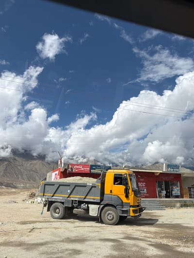 A bright yellow and gray dump truck, laden with a light-colored aggregate, is parked on a dirt surface in front of a row of shops. The truck has its headlights on, and a person is visible in the driver's seat, looking forward. The shops have red facades and display various products. A Coca-Cola advertisement featuring a woman drinking from a bottle is prominently displayed on one of the shop fronts. Behind the shops, rugged, barren mountains rise under a sky filled with dramatic, fluffy white clouds against a deep blue expanse. Power lines are strung across the scene, adding to the sense of a remote, possibly mountainous, location. The scene appears to be a roadside in Mankang, India, a town known for its mountainous terrain and proximity to tourist attractions.
