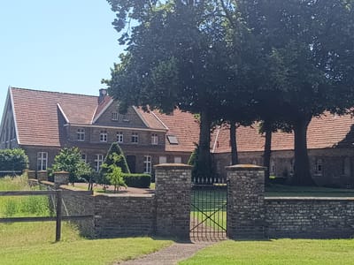 The image shows a large, historic brick building with a red tile roof, situated in a rural or semi-rural setting. The building appears to be a manor house or a complex of farm buildings. It features multiple windows with white frames and several chimneys. The architecture suggests it might be of European origin. Lush green trees with dense foliage are prominent in the foreground, partially obscuring the building and casting shadows. A brick wall with stone pillars surrounds the property, and a black metal gate is visible in the center. A dirt path leads to the gate. The ground is covered in green grass, and in the background, there's more greenery and clear blue sky. The scene is peaceful and serene, suggesting a sunny day. The building is identified as a location in Telgte, Germany.