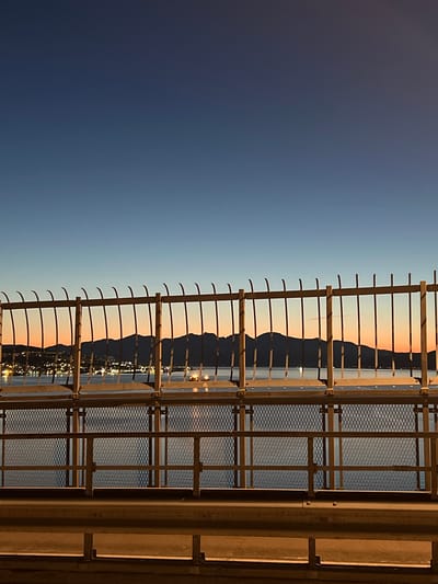 The image displays a serene coastal landscape at twilight, viewed from behind a prominent metallic structure. The foreground, occupying the right side of the frame, features a robust barrier or railing, possibly part of a bridge or viewpoint. This structure consists of thick vertical and horizontal metal bars, some sections integrated with a diamond-patterned mesh. It is warmly illuminated with a golden-brown light, creating a stark contrast with the cooler tones of the background.

Beyond the metallic barrier, a dramatic silhouette of a mountainous or hilly coastline extends across the midground. The dark, rugged peaks are sharply outlined against the luminous sky. At the base of the mountains, numerous small, bright lights twinkle, reflecting on a body of water, likely a fjord or sea. These lights suggest a coastal city or settlement.

The sky, filling the left and upper portions of the image, exhibits a striking twilight gradient. It transitions from a deep, dark blue at the top, gradually fading into a lighter, softer blue, and then to a warm, ethereal orange-pink hue just above the mountain peaks. This rich color palette indicates either dawn or dusk, with artificial lights already active. The weather appears clear with no visible clouds. The scene conveys a sense of peaceful grandeur, typical of the natural landscapes found in locations like Tromsø, Norway. No specific activity or visible text is present in the image.