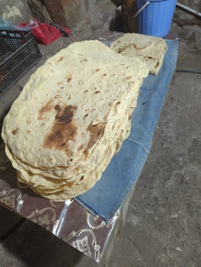A stack of flatbreads rests on a blue fabric, likely denim, on a patterned surface. The flatbreads are pale yellow with irregular brown, charred spots, suggesting they were baked. To the left, a black plastic crate and a red plastic bag are visible, with a hint of a blue bucket in the background to the right. The overall scene appears to be outdoors or in a rustic, unfinished indoor space, with a concrete floor and rough stone or brick walls. The lighting is dim. The location is the city of Solak, Armenia.