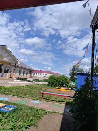 The image depicts a sunny day with a blue sky dotted with white clouds. In the foreground, there is a blue play structure with a green fence. A string of colorful triangular flags is strung across the sky, attached to a pole on the right. To the left of the play structure is a yellow sandbox. Further left, a red and blue bench sits on a path marked with colored circles. A blue bin filled with colorful plastic caps is on the ground near the bench. In the background, a school building with a light-colored facade and a red roof is visible. Another building with a red roof and white walls is also present in the distance. Lush green bushes and trees surround the buildings and playground area. The scene is set in the city of Nizhnyaya Makhtama, Russia, suggesting it is a playground or outdoor recreational area.