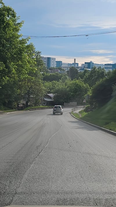 A light-colored SUV is driving away from the camera on a paved road. The road curves to the right. On the left side of the road, there are trees and some buildings. On the right side of the road, there is a grassy slope with a guardrail. In the distance, there are several modern buildings with blue and white facades, likely part of an industrial or institutional complex. The sky is blue with some clouds. The scene appears to be in an urban or suburban area. The weather is clear and sunny.