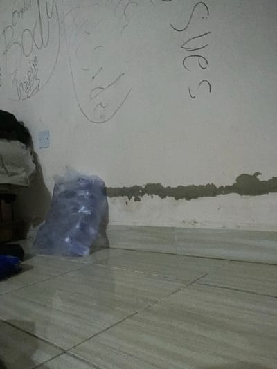 The image depicts a close-up, low-angle view of a textured wall with writing and a floor. On the wall, there are several pieces of writing in what appears to be pencil or charcoal. To the left, the word "Body" is written in large, stylized letters, with "Inspire" written below it in a smaller script. To the right of "Body," there is a sketch of a face, with an eye, nose, and mouth depicted. Further to the right, the word "Sves" is written vertically. The wall itself has a rough texture and a dark, uneven horizontal line running across it, suggesting damage or a change in material.

In the foreground, on the floor, there is a clear plastic bag filled with what appears to be multiple plastic bottles. The bag is translucent, and some of the bottles have a slight purple hue. The floor is tiled with light-colored square tiles, and grout lines are visible. To the left of the bag, a dark object, possibly clothing or a bag, is partially visible. A white electrical outlet is mounted on the wall above and to the left of the plastic bag.

The scene appears to be an indoor environment, likely a room. The lighting is dim, contributing to the grainy appearance of the image. The overall impression is one of a somewhat unkempt or unfinished space.

This scene is located in Gbaza, Nigeria.