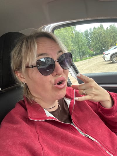 A blonde woman wearing sunglasses and a red zippered jacket is sitting in the driver's seat of a car. She is looking at the camera and making a "call me" gesture with her right hand. The car appears to be parked outdoors, with trees and a white car visible in the background. The weather appears to be overcast. The location is identified as Chastyye, Russia based on a visible license plate in the background that reads "CHASTYE" and "37 RUS".