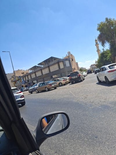 The image captures an urban street scene in Amman, Jordan, specifically in the vicinity of Abdoun Circle (Dawar Abdoun). The viewpoint is from inside a stationary or slowly moving vehicle, looking forward and slightly right.
In the immediate foreground, the dark frame of a car's interior is visible on the left, along with a side mirror reflecting a portion of a light-colored vehicle and the arm of an individual wearing a blue sleeve. The asphalt road surface, appearing dry, extends through the midground.
Multiple vehicles are present on the road and parked along the curb. A white sedan is partially visible in the left midground, oriented with its rear towards the viewer. Further into the midground, a gold/beige sedan is prominent among several other parked or moving cars of various colors.
The background is dominated by urban architecture. On the left, a multi-story building with a light-colored facade features the word "FARMASHOP" prominently displayed. This building has a distinctive dark, possibly metallic, structure on its roofline. To the right of this building, a tall, slender, light-colored mosque minaret with a pointed cap rises into the sky, partially framed by green tree foliage. A black street lamp post is visible on the far left.
The sky is clear and bright blue, indicating sunny weather conditions. No distinct human figures are visible beyond the partial reflection in the side mirror.