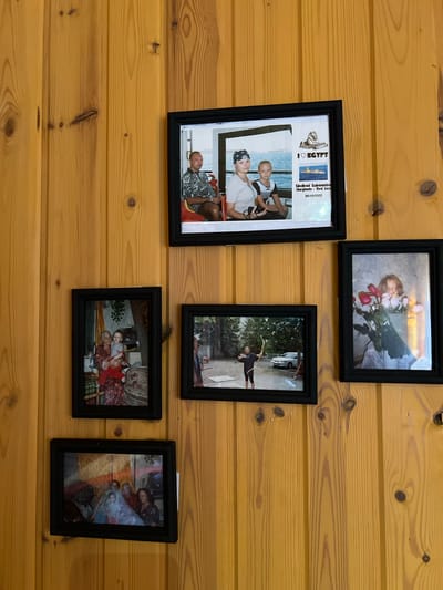 The image displays a collection of framed photographs hanging on a warm-toned, vertically paneled wooden wall. The overall aesthetic suggests a domestic setting, possibly a home in Трасна Снт, Russia, given the context.
The top photograph, framed in black, shows a woman wearing a headscarf and a young boy posing for a picture. Behind them is a body of water and a distant shoreline, suggesting a vacation or travel. To the right of this picture, a text overlay reads "I LOVE EGYPT" with an image of the Sphinx and a submarine.
Below this, on the left, is another framed photograph of an elderly woman holding a young child. Both are smiling and dressed in colorful clothing. The background appears to be an interior, possibly a living room with patterned curtains.
To the right of the previous photo, and slightly lower, is a framed picture of a young girl with auburn hair, holding a bouquet of pink roses. The lighting in this image is soft, highlighting the girl's face and the flowers.
Directly below the picture with the elderly woman and child, a framed photograph shows a group of people, possibly a family, sitting together. Their expressions are gentle, and the ambiance is familial.
On the far right, and slightly above the photograph of the girl with roses, is a framed image of a man seated in a red chair, wearing a patterned shirt. He looks towards the camera with a calm expression. The background appears to be a window with a view of water.
In the lower left quadrant, another framed photograph depicts a person aiming a bow and arrow in an outdoor setting. A car is visible in the background, suggesting a casual activity or hobby.