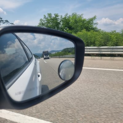 The image is a photograph taken from inside a moving vehicle, looking into the side-view mirror. The mirror reflects a highway scene in Ucheon-myeon, South Korea, under a bright, cloudy sky. The primary subject in the mirror is a dark-colored truck driving on the highway, appearing to be in the lane adjacent to the photographer's vehicle. The highway itself is paved and has white lane markings. A metal guardrail runs along the side of the highway, separating it from a grassy embankment and lush green trees and vegetation. In the distance, rolling hills covered in trees are visible. The reflection in the mirror also shows the sky with scattered white clouds. The side of the photographer's white car is visible in the foreground, partially obscuring the reflection in the mirror. A small, circular convex mirror is attached to the main side-view mirror.