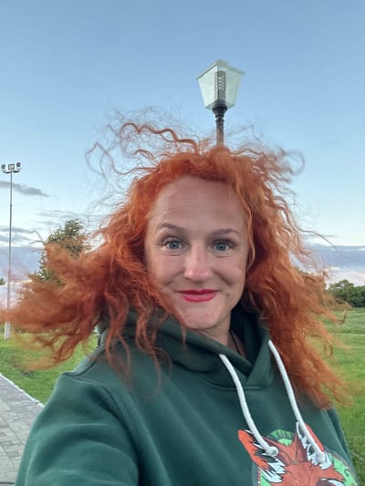 A woman with bright red, curly hair that is being blown by the wind is captured in a selfie. She is wearing a dark green hooded sweatshirt with a graphic design of a fox's head on the front. Her lips are painted with bright red lipstick, and she is smiling at the camera. In the background, a vintage-style street lamp with a hexagonal glass shade stands tall against a pale blue sky with scattered clouds. To the left, a tall light pole with multiple spotlights is visible, along with a paved walkway and a grassy field. In the distance, trees can be seen on the horizon. The overall scene suggests an outdoor setting, possibly a park or a public space in the city of Bolgar, Russia, during either dawn or dusk, given the lighting conditions.