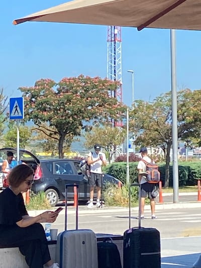 In Golubovci, Montenegro, on a bright, clear day, a group of individuals are gathered outdoors, likely at a transportation hub or waiting area. A woman in a black dress and sunglasses sits on a bench, engrossed in her mobile phone. Beside her are two suitcases. A few feet away, a man in a white t-shirt, black shorts, and a cap stands by a car with its trunk open, also looking at his phone. Another man in a similar outfit, carrying a backpack, is seen in the mid-ground. In the background, a tall, red and white lattice tower pierces the clear blue sky. Lush green trees with pink blossoms line the area, and a pedestrian crossing is visible in the foreground, marked by orange cones. The scene suggests a moment of transit, with people waiting and engaged with their devices before continuing their journeys.