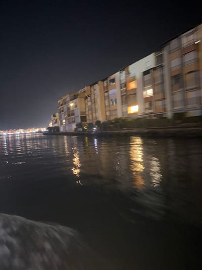 This is a nighttime, medium-shot, slightly blurry photograph taken from a body of water, likely a canal or river, looking towards a waterfront of buildings and the sky. The water in the foreground is dark and reflects the lights from the buildings, creating shimmering streaks of orange, yellow, and white. A white wake is visible in the lower left corner, indicating movement.
The buildings are a mix of multi-story structures with balconies and windows, illuminated from within. The architecture appears modern, with light-colored facades and various window designs, some with closed shutters. The buildings stretch along the waterfront, receding into the distance on the left where more lights are visible, suggesting a populated area. The sky above is dark with a few faint stars visible.
The scene is located in Agde, France, likely along the Hérault River or the Canal du Midi, given the presence of waterfront buildings and the reflective water. The overall impression is one of a tranquil, illuminated waterfront at night. There are no people clearly visible in the image.