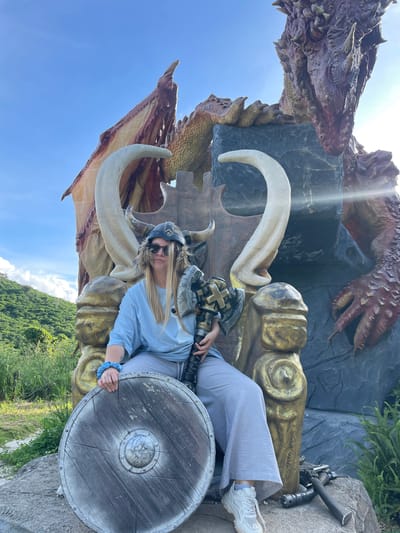 A person sits on a throne in front of a dragon statue. The person is wearing a Viking helmet with fur trim, sunglasses, a blue t-shirt, and grey pants. They are holding a large axe in their right hand and a round shield on their left. The throne is made of gold-colored material and appears to be carved to resemble tusks and possibly a rough, rocky texture. The dragon statue is large and reddish-brown with prominent scales and wings. Behind the throne, a rocky structure is visible, with a portion of the dragon's body and head appearing to emerge from it. The background shows a bright blue sky with some scattered clouds, lush green hills covered in vegetation, and some smaller green plants in the foreground. The overall scene suggests an outdoor attraction or themed environment, possibly a tourist spot. No specific location within Thôn 1, Vietnam is identifiable from the image alone.