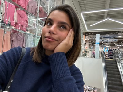 A woman is depicted in a retail setting identified as a Primark store, specifically located at 1001 Market Street in Philadelphia, Pennsylvania, United States. She has medium-toned skin, dark, shoulder-length hair, and is wearing a navy blue, textured long-sleeved sweater with a black strap from a shoulder bag visible. Her right hand rests against her jawline as she looks upwards and to the right with a pensive expression. To her left, behind clear display panels, multiple rows of children's clothing, predominantly in pink, purple, and grey hues, are visible. To her right, a downward-moving escalator is present, leading to a lower retail floor where various merchandise displays and distant shoppers can be observed. The ceiling features geometric fluorescent lighting, including square and triangular configurations. A vertical sign column indicating "2" is visible, and a reversed reflection in the background displays the word "PRIMARK."